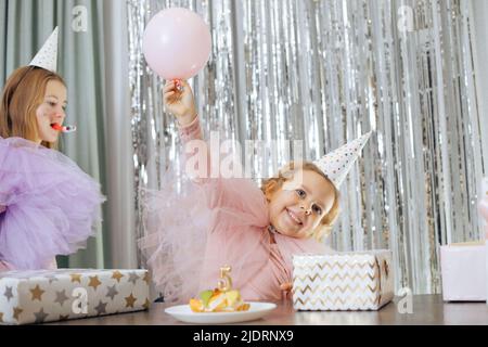 Magnifique anniversaire. Très joyeux bébé de 5 ans dans le chapeau de fête a soulevé sa main avec le ballon devant elle est gâteau d'anniversaire et beaucoup de boîtes avec des cadeaux Banque D'Images