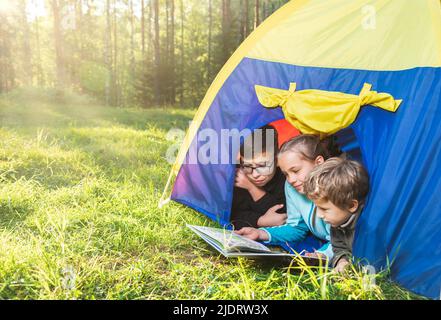 Les enfants lisant un livre allongé ensemble dans une tente sur un pré ensoleillé appréciant le camping de la forêt d'été. Le soleil brille à travers les arbres. Familiale été vac Banque D'Images