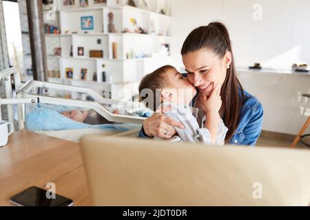 Garçon donnant à la mère un baiser sur la joue au PC dans le salon avec bébé en arrière-plan Banque D'Images