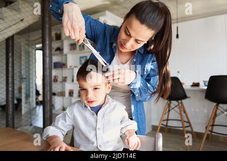 La mère ou le coiffeur coupent les cheveux d'un garçon avec un peigne et des ciseaux dans le salon de coiffure Banque D'Images
