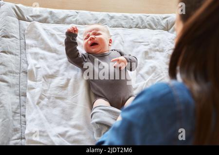 Mère met un costume de barboteuse sur un bébé qui pleure après avoir changé de couches Banque D'Images