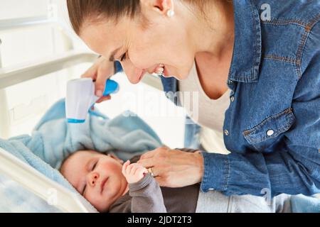 La mère soignante mesure la fièvre du bébé au lit à l'aide d'un thermomètre frontal sans contact Banque D'Images