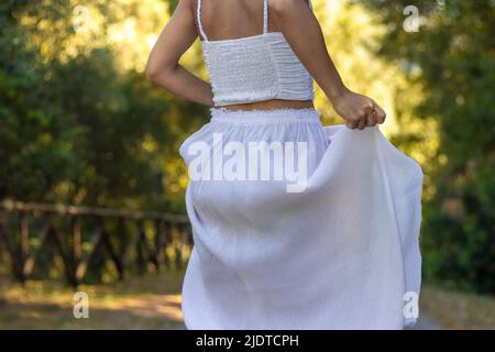 Vue coupée d'une jeune femme méconnaissable se déplaçant et tenant sa longue jupe boho blanche avec la nature en arrière-plan Banque D'Images