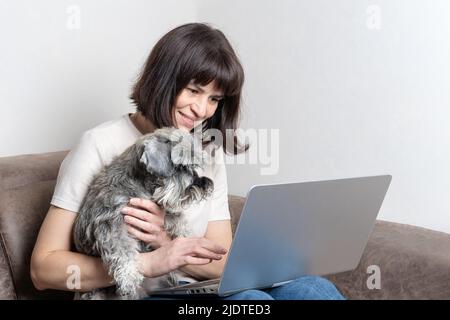 Belle femme d'âge moyen avec un adorable chien schnauzer miniature travaille à la maison sur un ordinateur portable tout en étant assis sur le canapé. Une femme d'âge moyen travaille à distance Banque D'Images