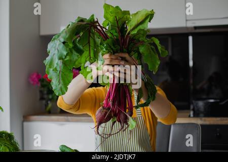 Femme blonde se cachant le visage derrière un bouquet de betteraves avec des feuilles, faire cuire la salade biologique fraîche dans la cuisine. Alimentation saine, alimentation Banque D'Images