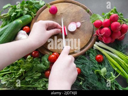 Les mains coupent des radis avec un couteau en céramique blanc sur une planche à découper ronde en bois. Autour de la carte sont des légumes frais mûrs: Tomates, oignons, cucu Banque D'Images