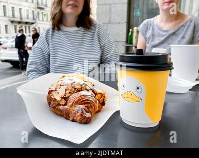 Saint-Pétersbourg, Russie, août 2021 : café dans une tasse en carton jaune avec croissant de canard et d'amande. Deux filles sur un fond flou. Banque D'Images