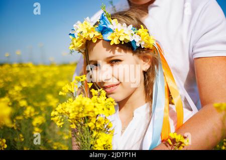 Frère câlins soeur avec couronne ukrainienne et bouquet de fleurs dans les mains, dans champ de colza sous ciel Banque D'Images