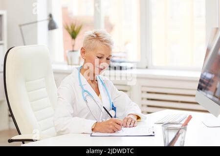 Souriant beau médecin mature avec stéthoscope assis au bureau et ajoutant des informations sur le patient dans le dossier médical tout en travaillant dans son propre bureau Banque D'Images