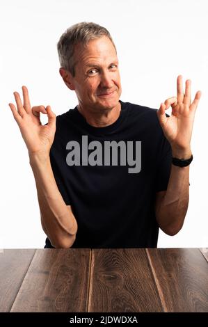 Un homme souriant, beau, âgé, dans un tee-shirt bleu avec les deux mains vers le haut, signalisation OK, sur fond blanc. Banque D'Images