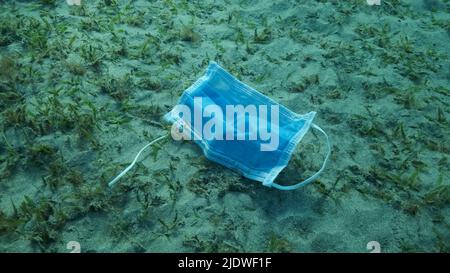 Le masque utilisé repose sur un fond sablonneux recouvert d'un parfum vert de senne sous la lumière du soleil. Les masques rejetés polluent les fonds marins depuis le coronavirus COVID-19. Polluti Banque D'Images