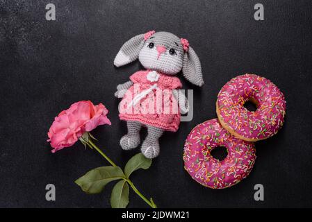 Magnifique jouet de lapin tricoté avec des beignets à la glaçure rose et saupoudrés de couleur sur un fond de béton foncé Banque D'Images