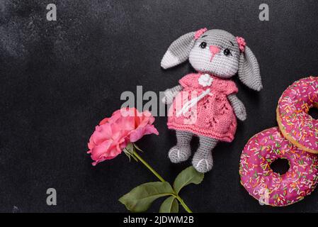 Magnifique jouet de lapin tricoté avec des beignets à la glaçure rose et saupoudrés de couleur sur un fond de béton foncé Banque D'Images