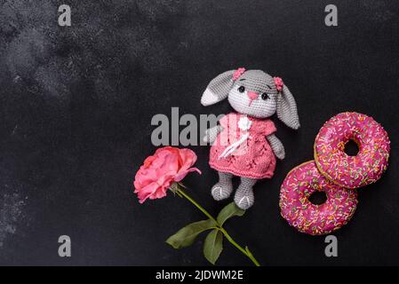 Magnifique jouet de lapin tricoté avec des beignets à la glaçure rose et saupoudrés de couleur sur un fond de béton foncé Banque D'Images