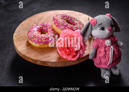 Magnifique jouet de lapin tricoté avec des beignets à la glaçure rose et saupoudrés de couleur sur un fond de béton foncé Banque D'Images