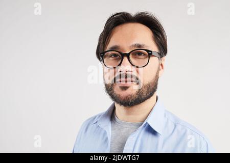 Portrait du contenu confiant jeune homme à barbe asiatique en lunettes debout sur fond gris clair Banque D'Images