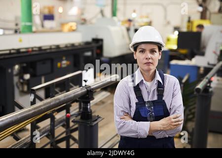 Portrait d'une femme expérimentée sérieuse et confiante travaillant dans une grande production de métaux, debout dans un atelier industriel et croisant les bras sur la poitrine Banque D'Images