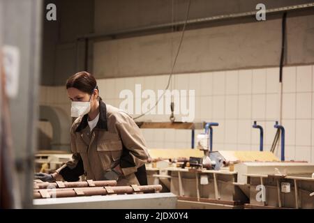 Femme chargée de brunette dans un respirateur debout dans un atelier industriel poussiéreux et examinant des tubes traités Banque D'Images