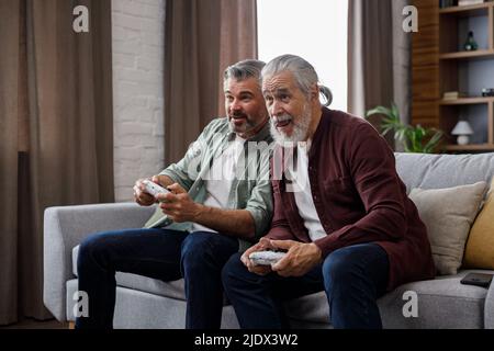 Le fils adulte et le père senior s'amusent, aiment passer du temps ensemble. Deux générations de famille jouant à des jeux vidéo en utilisant des gamepads à la maison. Fête des pères Banque D'Images