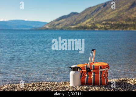 Pique-nique d'été sur une plage. Thermos et dos avec nourriture sur la rive rocailleuse du lac Okanagan en début de matinée d'été sur le fond avec les montagnes. Banque D'Images