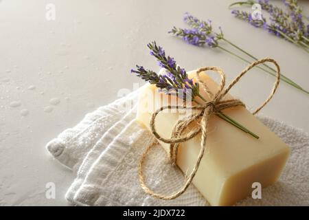 Détail de savon de bar naturel avec extrait de lavande sur serviette sur table blanche avec gouttes d'eau et pointes de lavande en fleur. Vue en hauteur. Horizonta Banque D'Images