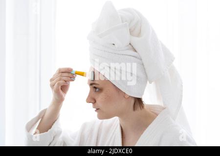 Jeune femme avec brosse appliquant un masque facial sur le front à la maison Banque D'Images