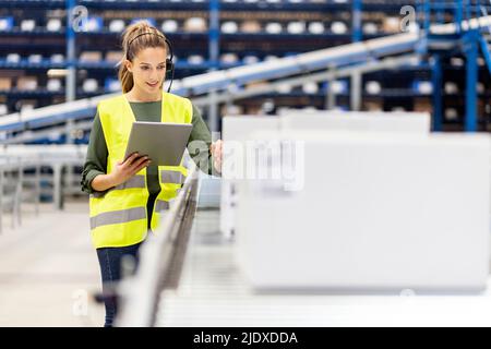 Jeune employé avec un PC tablette examinant des boîtes sur le tapis transporteur dans l'entrepôt Banque D'Images