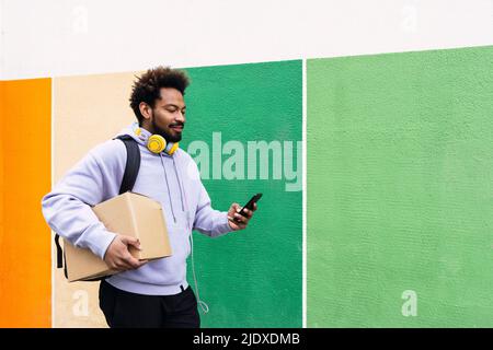 Le liveur tient la boîte à l'aide d'un smartphone devant un mur coloré Banque D'Images