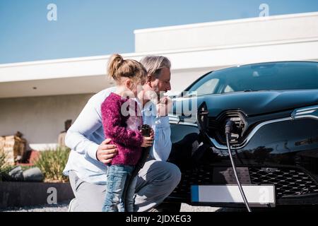 Fille tenant une fiche électrique debout par le père regardant la voiture le jour ensoleillé Banque D'Images