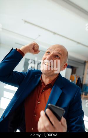 Un homme d'affaires enthousiaste tenant un téléphone intelligent pour faire des gestes au bureau Banque D'Images