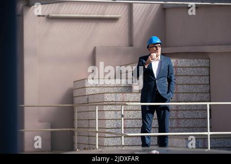 Architecte portant un casque de sécurité tenant une tasse jetable debout avec la main dans la poche sur le chantier de construction Banque D'Images