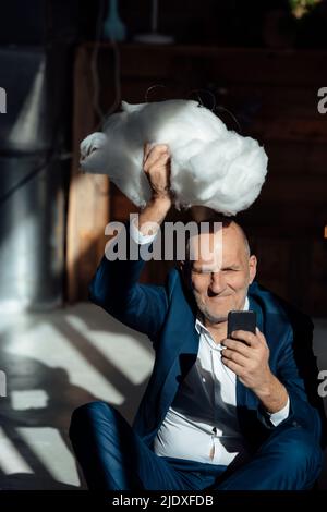 Homme d'affaires souriant tenant un téléphone mobile transportant le cloud au-dessus de la tête dans le bureau Banque D'Images