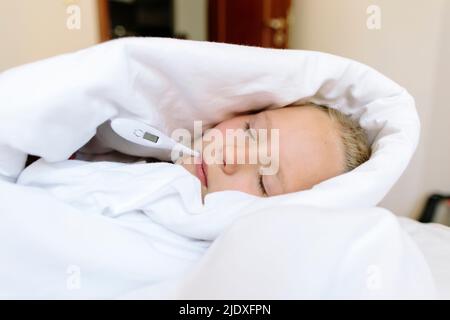 Fille malade enveloppée dans une couverture avec thermomètre sur le lit à la maison Banque D'Images