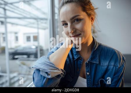 Femme souriante portant une chemise en denim assise près de la fenêtre à la maison Banque D'Images