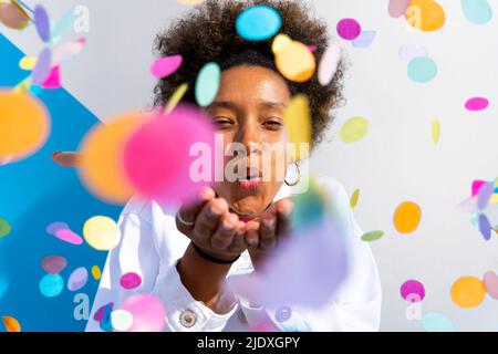 Jeune femme soufflant des confettis multicolores devant le mur le jour ensoleillé Banque D'Images