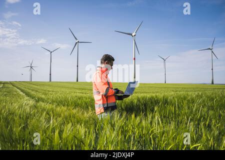 Ingénieur utilisant un ordinateur portable au milieu de récoltes de blé près des éoliennes sur le terrain Banque D'Images