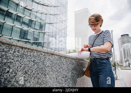 Femme d'affaires qui vérifie l'heure sur une montre intelligente près du mur de la ville Banque D'Images