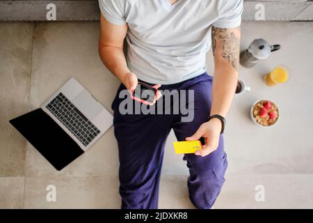 Jeune homme tenant une carte de crédit faisant des achats en ligne par téléphone intelligent à la maison Banque D'Images