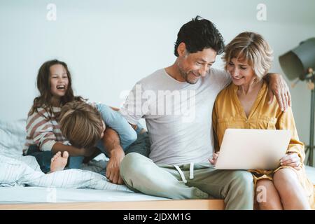 Un homme et une femme heureux avec un ordinateur portable assis par une fille et un fils jouant sur le lit à la maison Banque D'Images