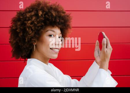 Jeune femme souriante tenant un miroir par un mur rouge Banque D'Images