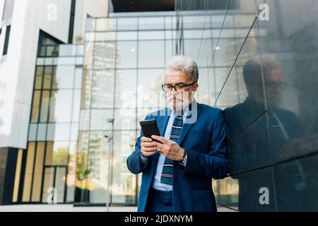 Homme d'affaires senior travaillant sur un smartphone à l'extérieur du bâtiment de bureau Banque D'Images