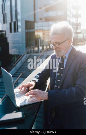 Homme d'affaires senior travaillant sur un ordinateur portable utilisant un chargeur de batterie solaire à l'extérieur du bâtiment de bureau Banque D'Images