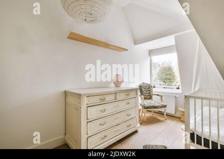 Intérieur d'une chambre moderne pour enfants avec une commode, un lit de bébé, une étagère, un fauteuil près de la fenêtre dans une chambre mansardée aux murs blancs Banque D'Images