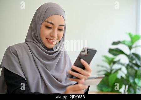 Jeune femme musulmane asiatique attrayante avec hijab utilisant un smartphone, discutant avec quelqu'un, défilant sur les médias sociaux, recherchant quelque chose sur le stagiaire Banque D'Images