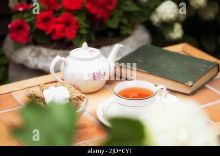 théière en porcelaine dans une tasse sur une table d'époque dans le jardin sur un fond de fleurs et un vieux livre en été Banque D'Images
