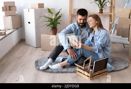 un jeune couple heureux s'assoit sur le sol dans des vêtements décontractés qui embrasent la photo encadrée. Une pile de boîtes en carton et de plantes de maison près d'un mur blanc à l'intérieur Banque D'Images