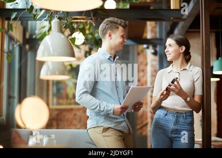 Des jeunes collègues satisfaits et positifs dans des vêtements décontractés se tenant dans un café confortable et utilisant un smartphone et un rapport d'analyse de marché tout en développant en ligne Banque D'Images