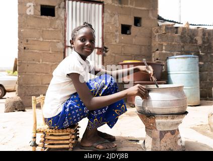 Petite fille africaine mignonne assise devant un four pour cuisiner de la nourriture pour sa famille dans une grande ville comme Bamako, au Mali Banque D'Images