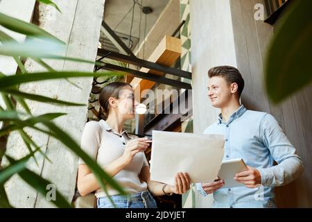Vue ci-dessous de jeunes créateurs positifs se tenant dans le hall contemporain et regardant les uns les autres tout en travaillant avec des croquis Banque D'Images
