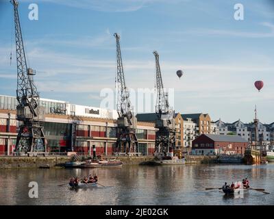 Des ballons à air chaud flottent au-dessus des grues, des bateaux à rames et du mShed dans la zone portuaire flottante de Bristol lors d'une soirée d'été ensoleillée. Banque D'Images